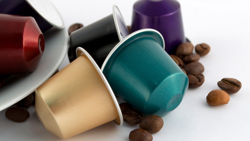 Plano de pormenor de seis cápsulas de café da Nespresso, cada uma de sua cor. Duas das cápsulas estão dentro de um prato (do qual só se vê parte da borda), e as restantes quatro estão à direita do prato. Ao redor destas quatro cápsulas são visíveis alguns grãos de café soltos.